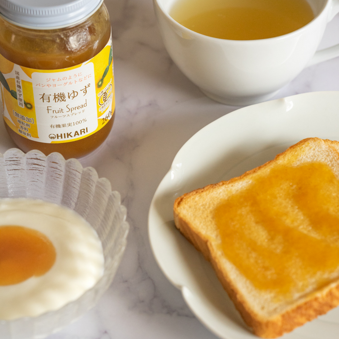A package bottle of the product surrounded by a slice of bread, a bowl of yogurt, and a cup of tea