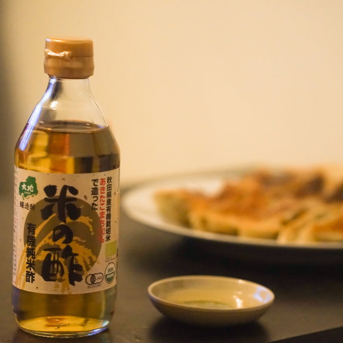 A bottle of the product and a small bowl of the organic rice vinegar in front of a plate of gyoza dumplings