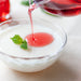 A bowl of yogurt being poured with rose syrup from glass sauce server
